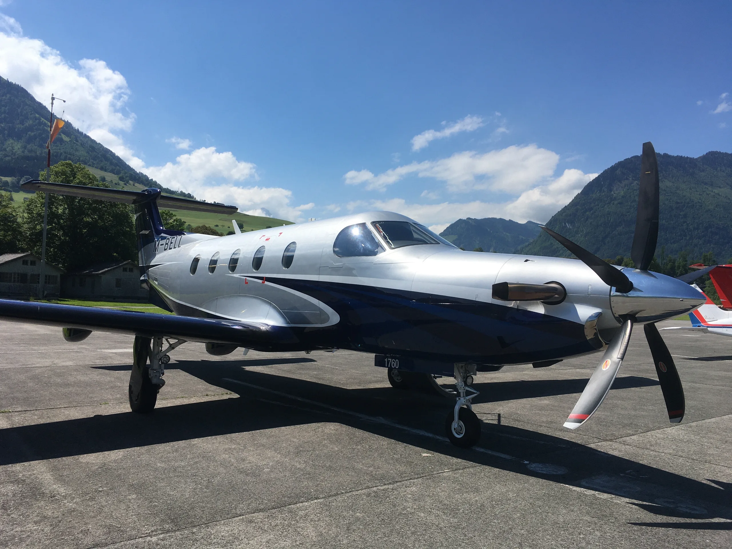 A silver and dark blue Pilatus PC-12 turboprop aircraft is parked on an airfield with mountains in the background.