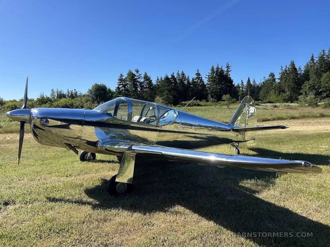 A shiny, chrome-like single-engine propeller aircraft with a bubble canopy is parked on a grassy field with trees in the background.