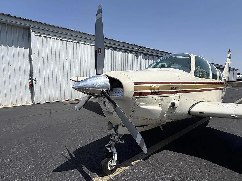 A close-up view of the front right side of a white and gold Beechcraft Bonanza, showing its propeller and nose.