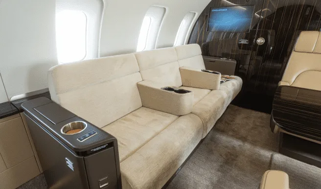 A beige sofa-style seat with cup holders and a side console inside a private jet cabin.