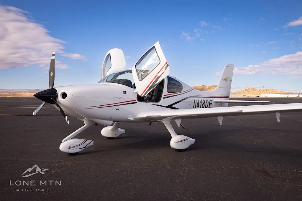 A white Cirrus SR22 aircraft with black and red stripes, registration N418DE, parked on an airfield with doors open.