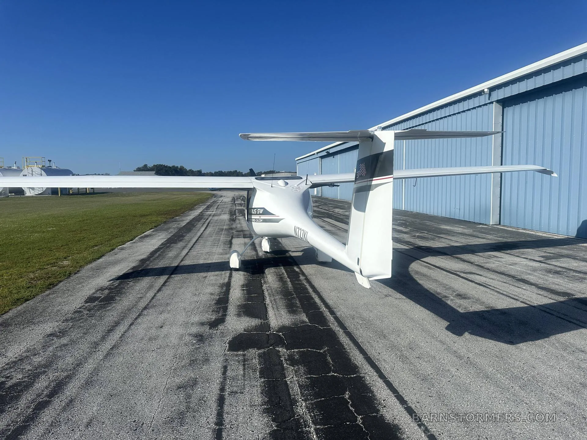 A white Pipistrel Virus SW aircraft is seen from the rear, parked on a tarmac in front of a blue hangar under a clear sky.