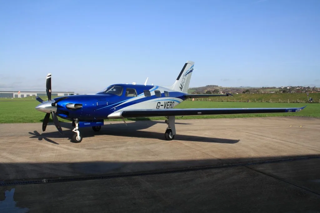 A three-quarter front view of a blue and silver Piper M600SLS aircraft (G-VERO) parked on an airfield tarmac.