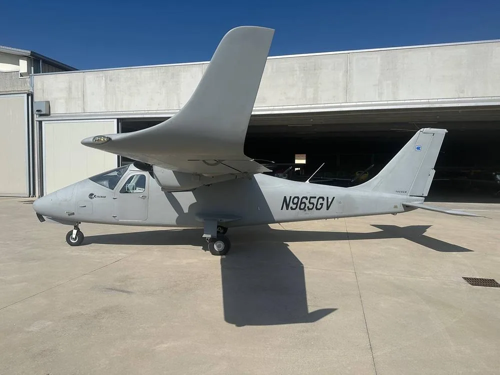 A light gray Tecnam P2012 Traveller aircraft with registration N965GV is parked on a tarmac in front of a hangar.