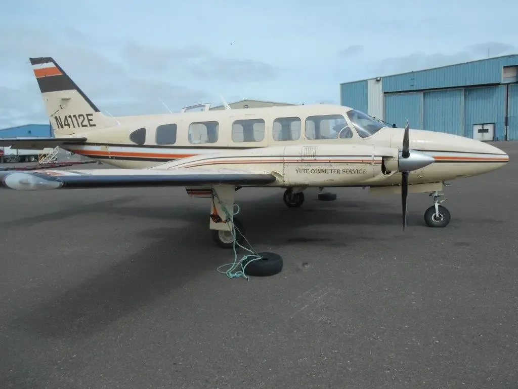 A beige twin-engine Piper Navajo aircraft, registration N4112E, with orange and black stripes, parked on an asphalt tarmac next to a blue hangar.
