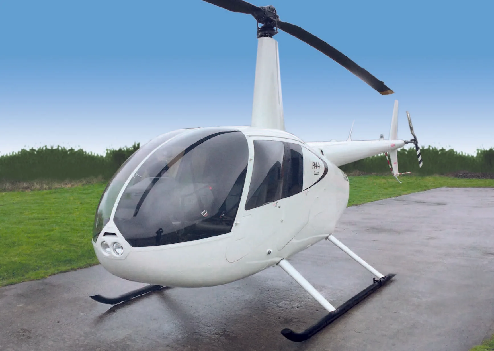 A white Robinson R44 Raven I helicopter is parked on a wet tarmac with a green and blue background.