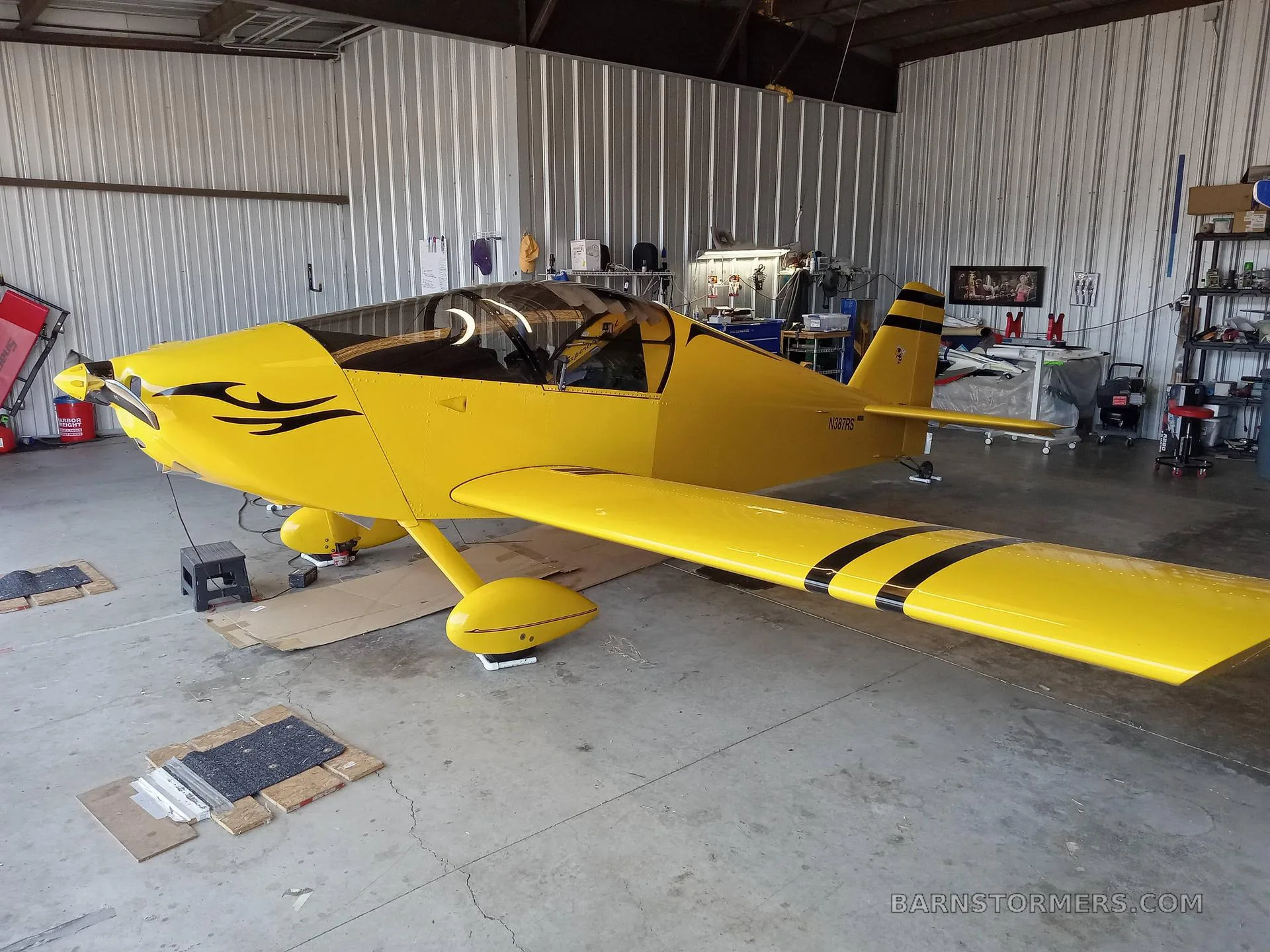 A bright yellow single-engine aircraft with black stripes and a black swirl decal on the nose, parked inside a hangar.