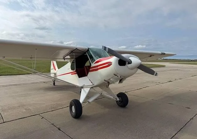A white and red single-engine taildragger aircraft is parked on a concrete tarmac under a cloudy sky.