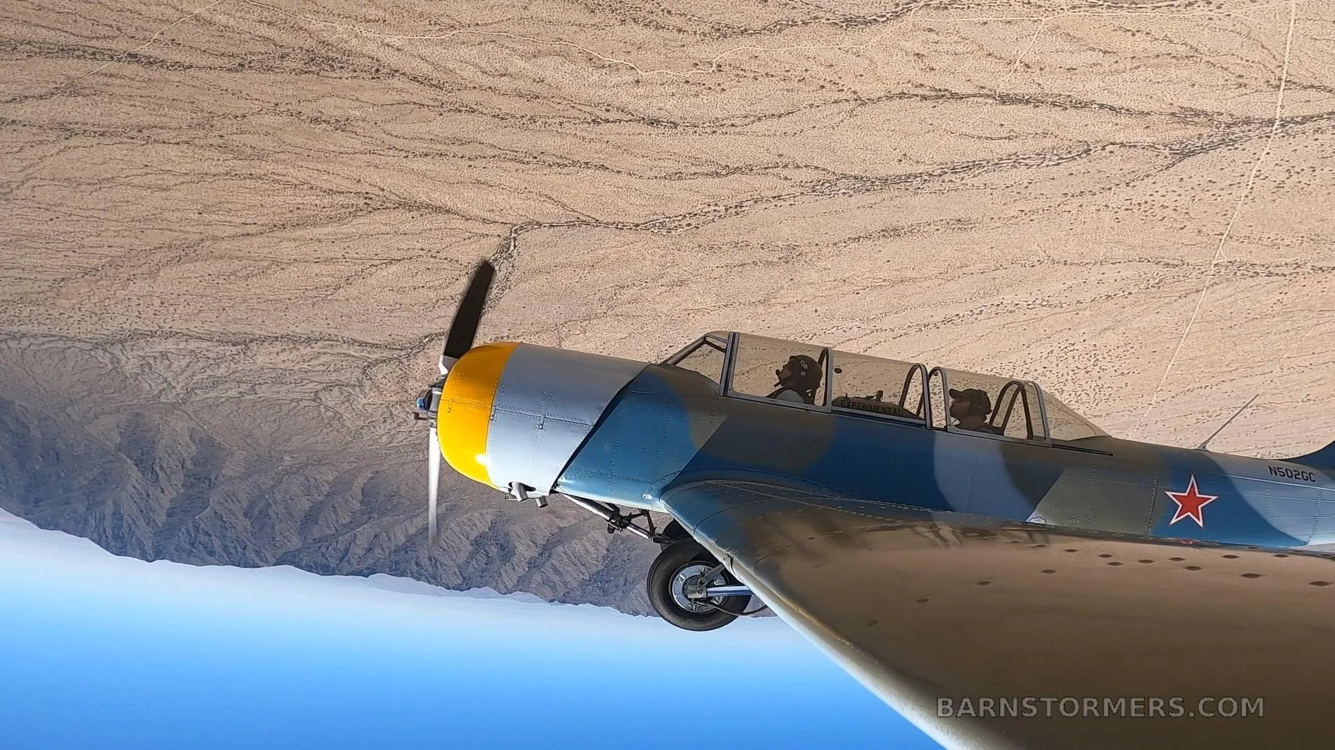 A blue and grey camouflage Yak-502GC aircraft with a yellow nose, seen from another aircraft, flying over a desert landscape.