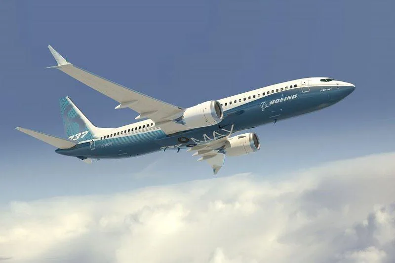 A Boeing 737 MAX in blue and white livery flying against a blue sky with some clouds.
