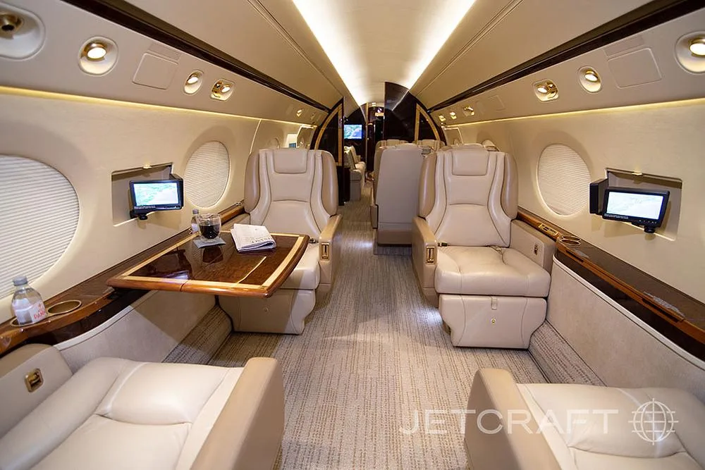 The luxurious interior cabin of a private jet, featuring beige leather seats, a wooden table, and ambient lighting.
