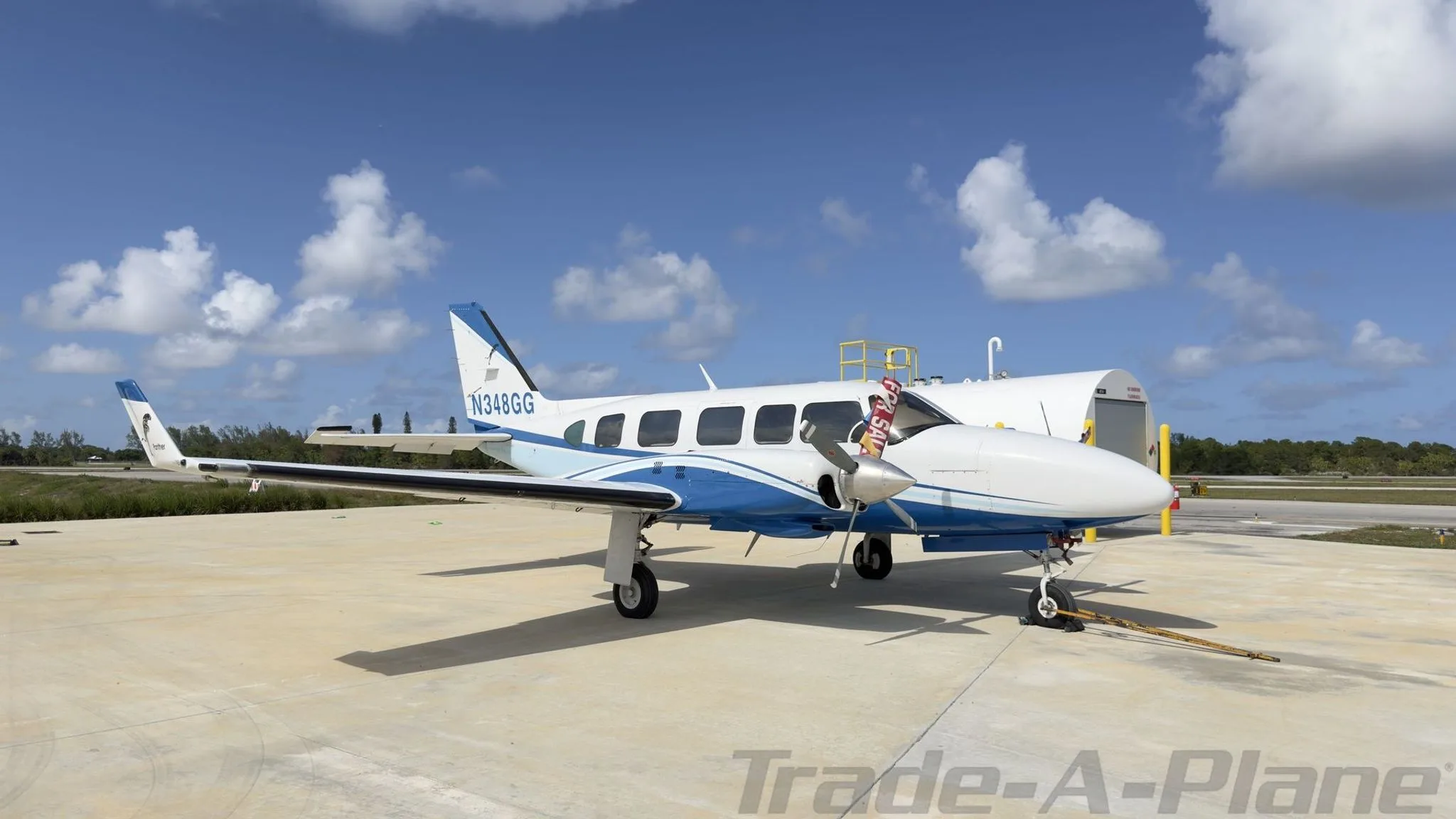 Side view of a white and blue twin-engine propeller aircraft, N348GG, with 'FOR SALE' tags on the propellers, parked on a concrete ramp.