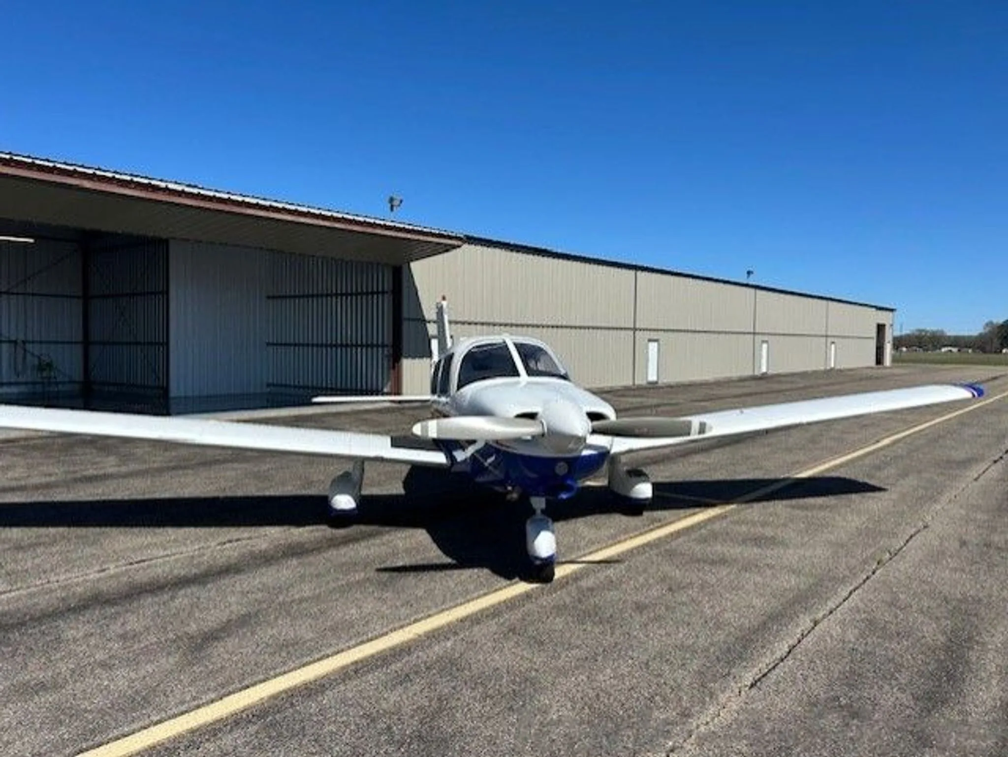 A front view of a white and blue single-engine aircraft parked on an airfield in front of a hangar.