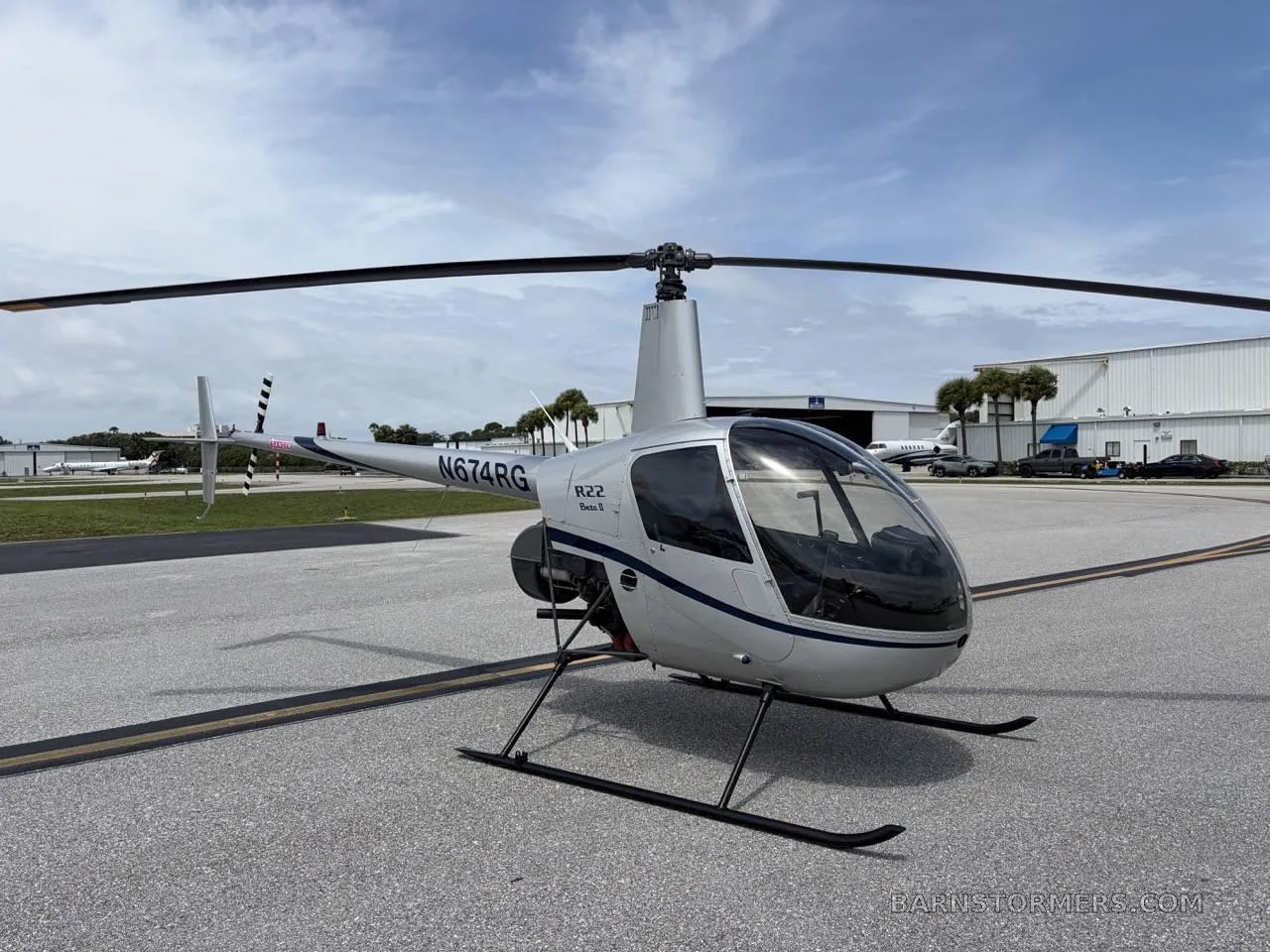 A silver Robinson R22 Beta II helicopter (N674RG) with blue stripes, parked on an asphalt tarmac at an airport under a partly cloudy sky.