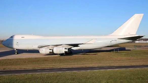 A large white Boeing 747 cargo aircraft with no livery is parked on an airfield under a clear sky.