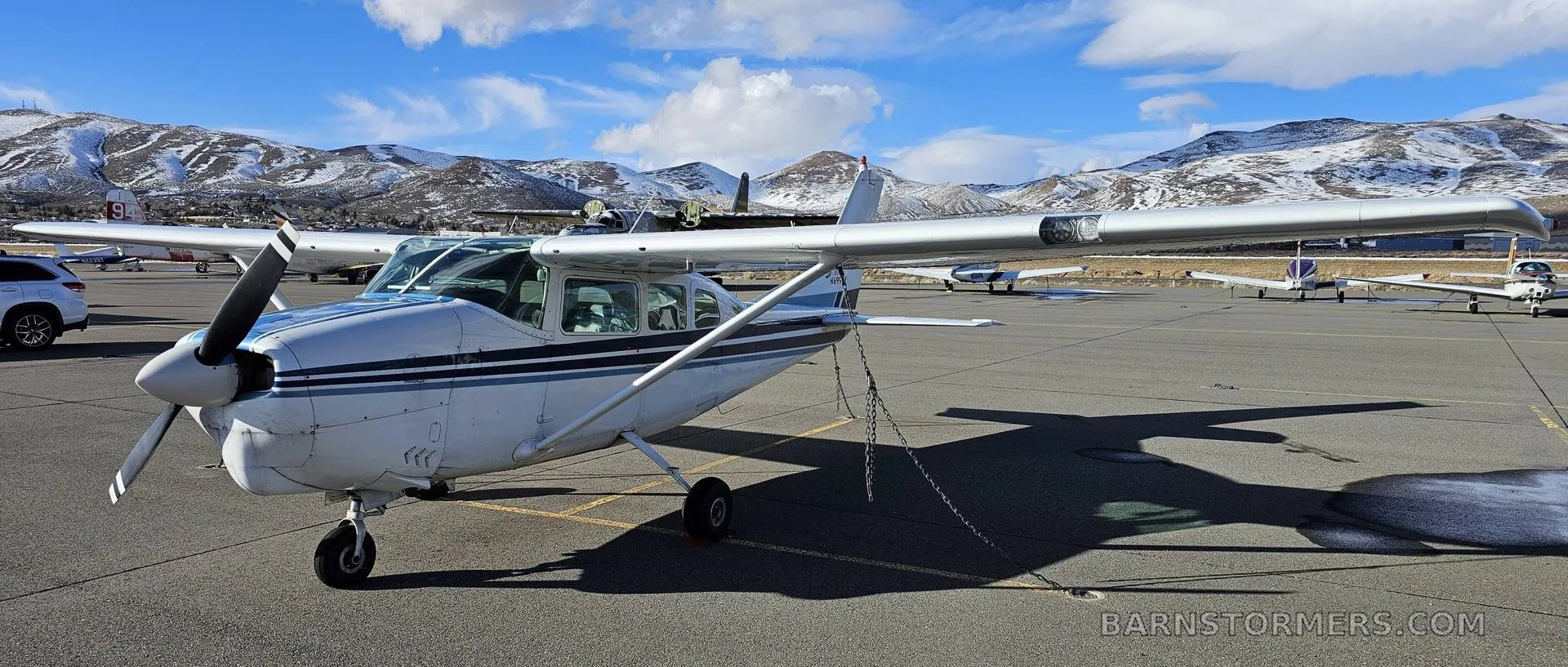 A white and blue Cessna 182 aircraft parked on an airfield with snowy mountains in the background.