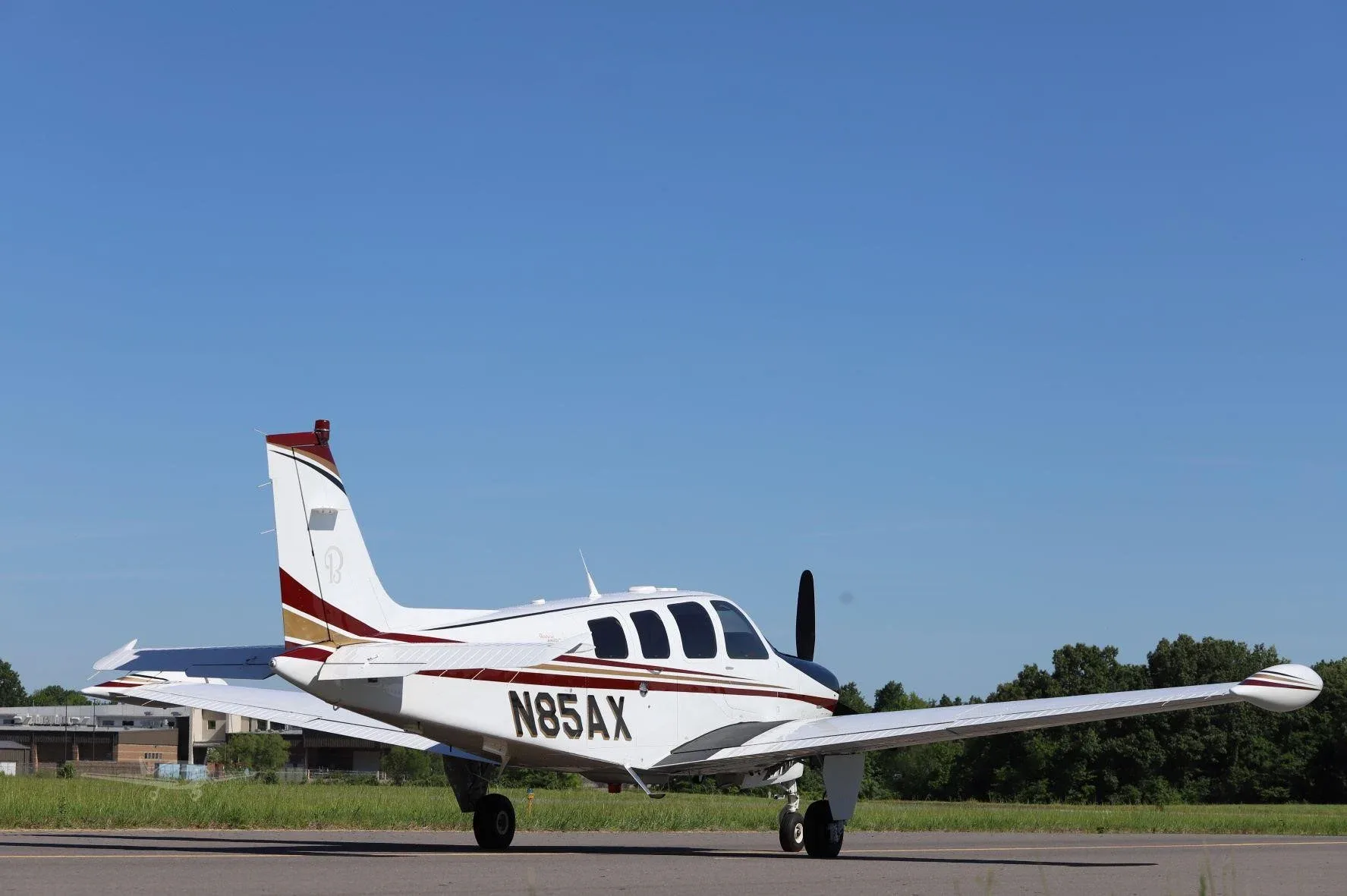 A white and black single-engine propeller aircraft with red and gold stripes and registration N85AX, viewed from the rear-right on a tarmac.
