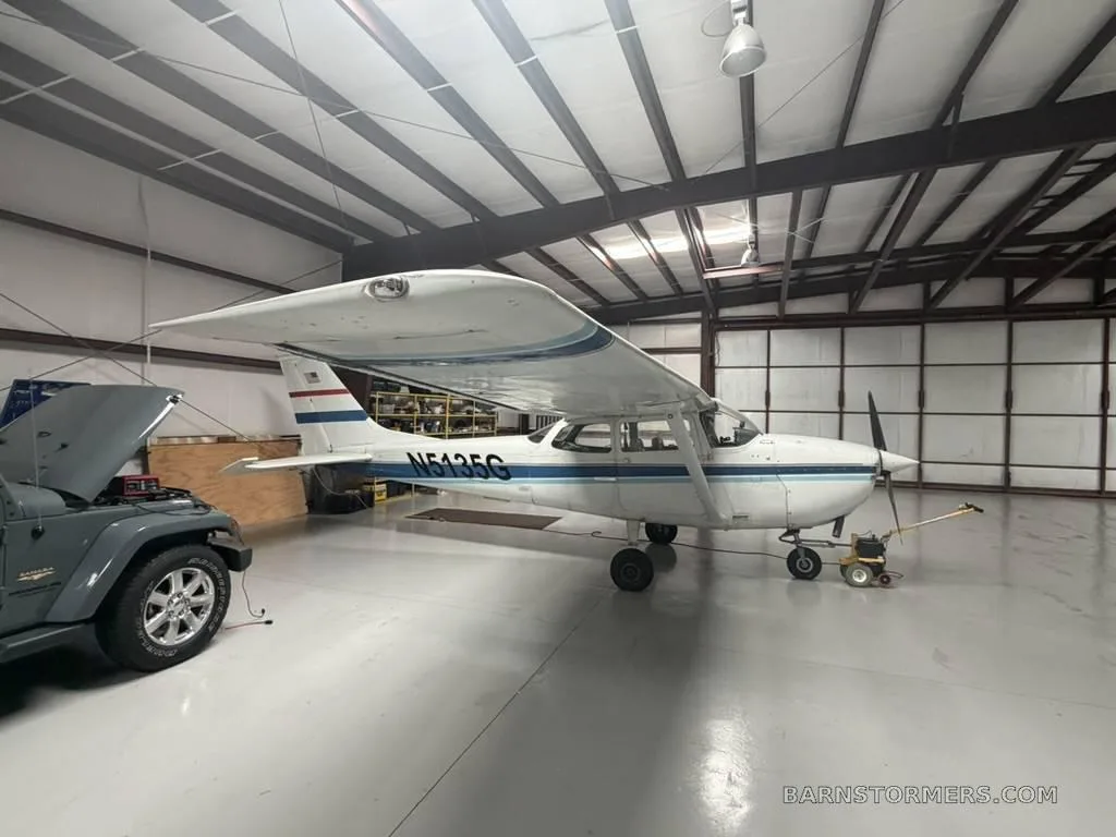 A white Cessna 172 (N5135G) with blue stripes is parked in a hangar next to a grey Jeep.