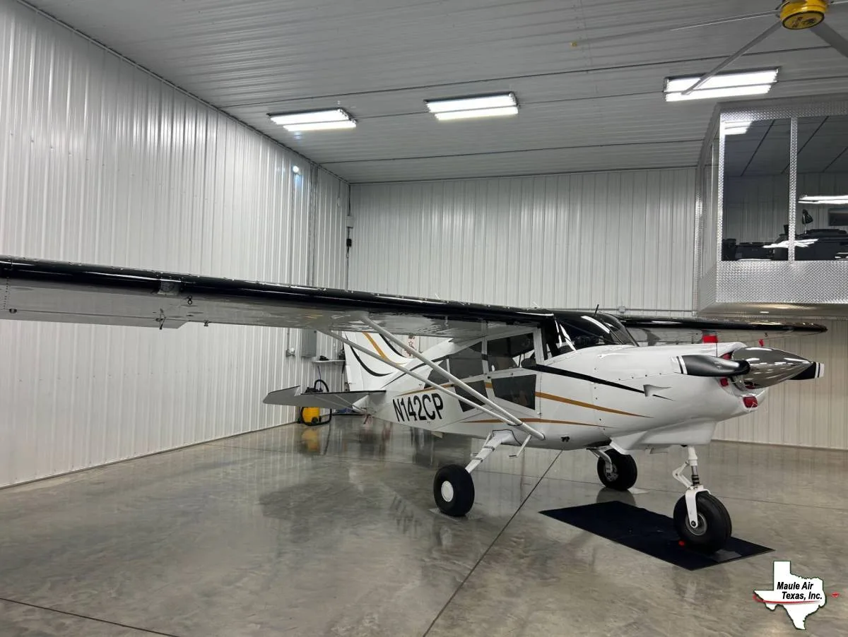 A white high-wing aircraft with black wings and gold stripes, featuring a nose wheel, sits in a hangar.