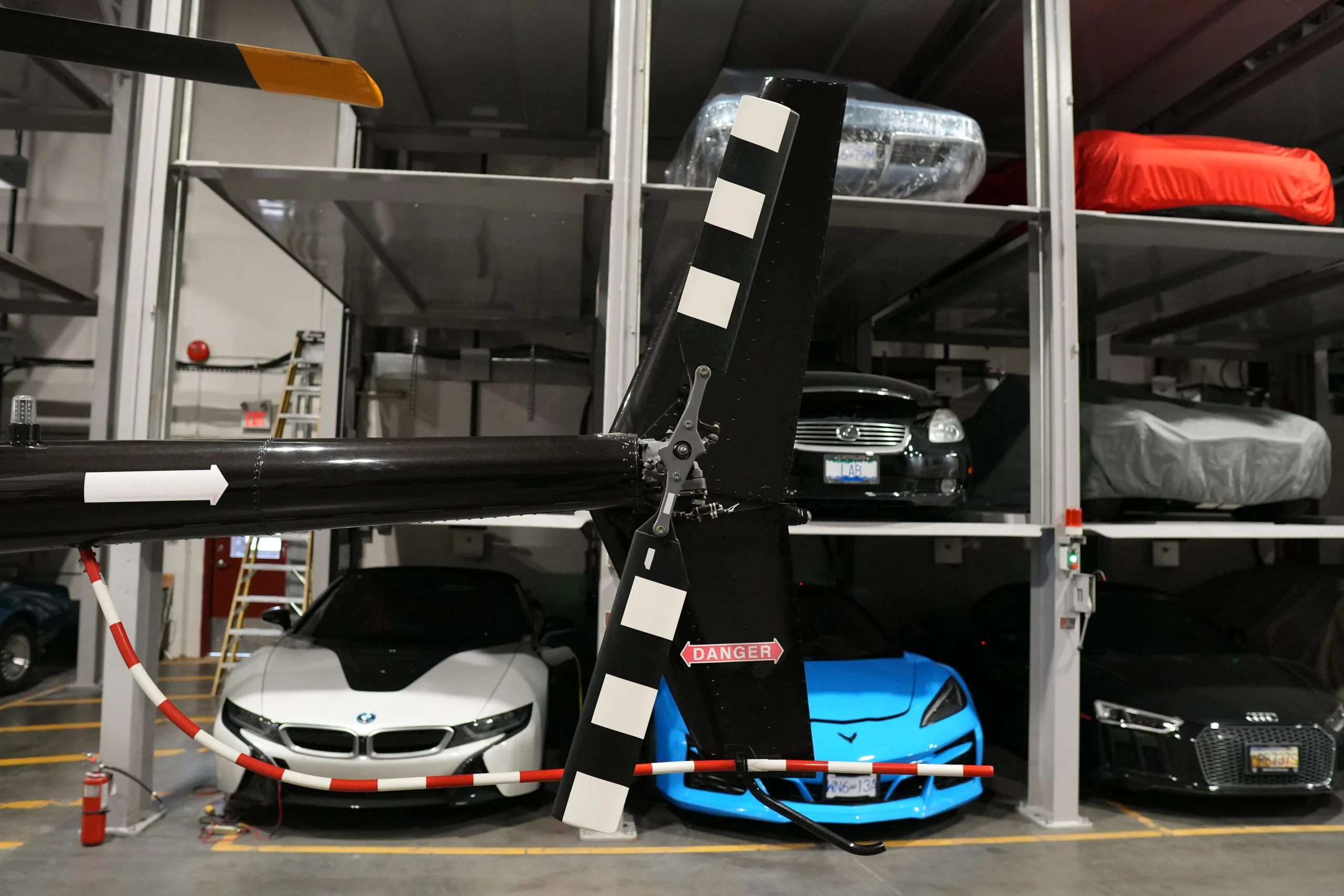 A close-up view of the black Robinson R44 helicopter's tail rotor with white stripes, surrounded by stored luxury cars.