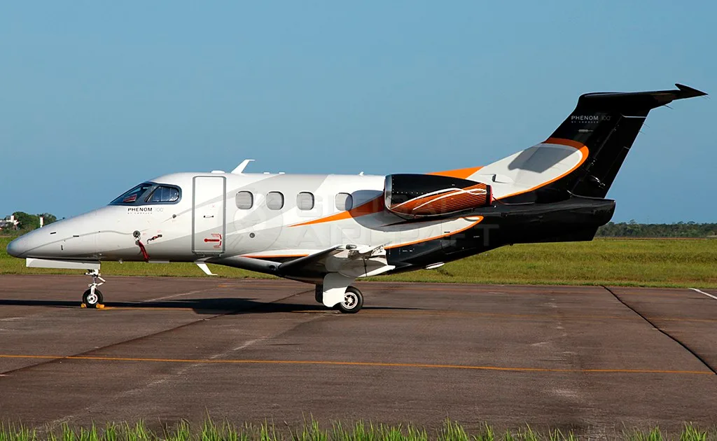 A white Embraer Phenom 100 business jet with black and orange accents is parked on a tarmac under a clear sky.