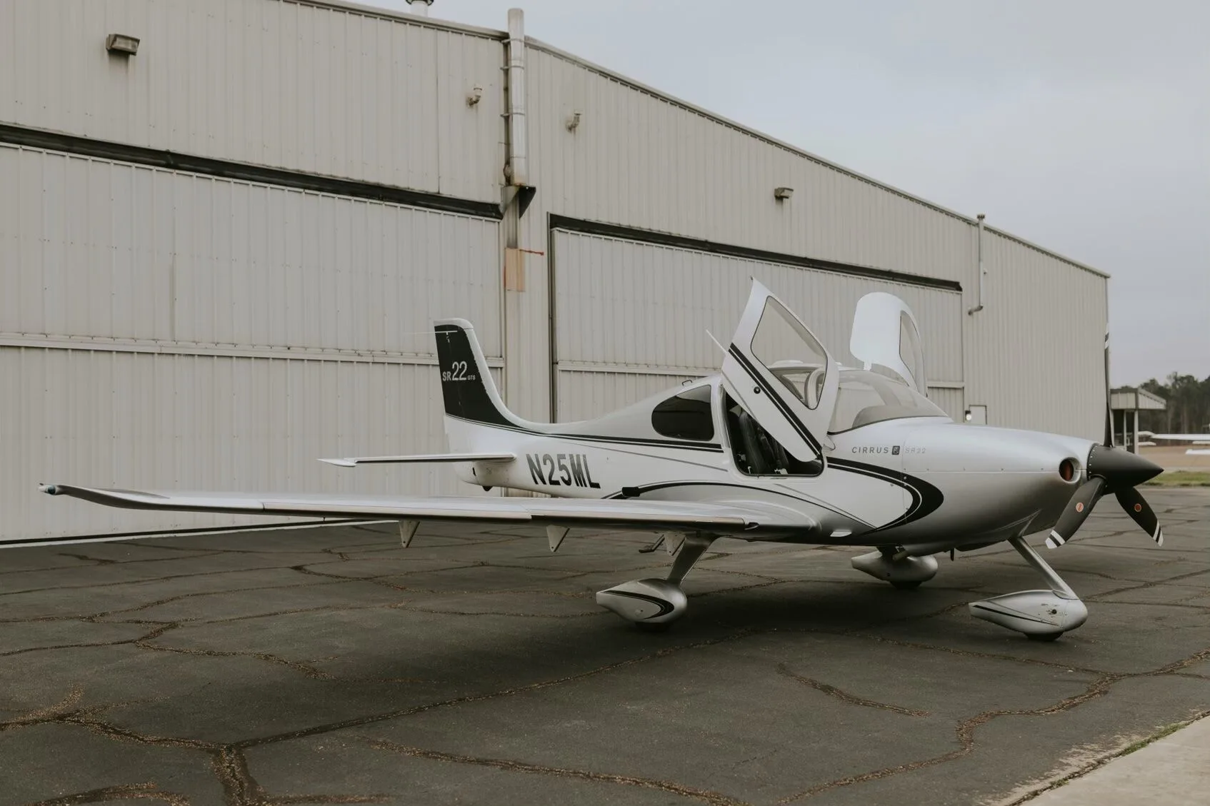 A silver Cirrus SR22 GTS aircraft with its gull-wing doors open, parked on an asphalt ramp in front of a hangar.