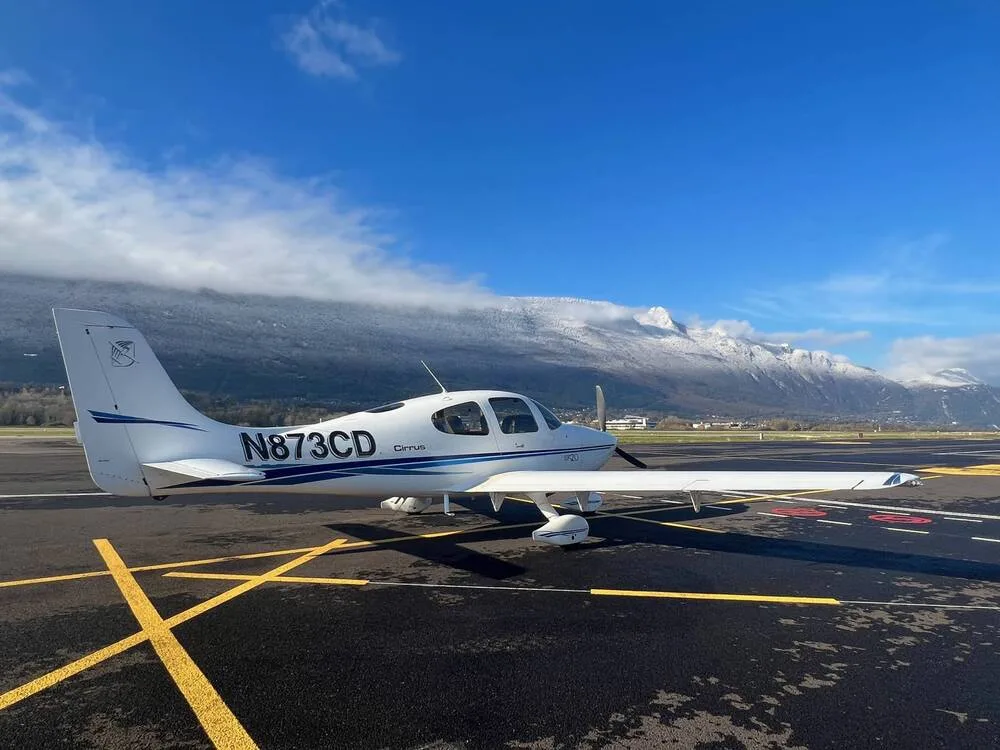 A white Cirrus SR20 aircraft (N873CD) with blue stripes parked on a wet tarmac, with snow-capped mountains in the background under a blue sky.