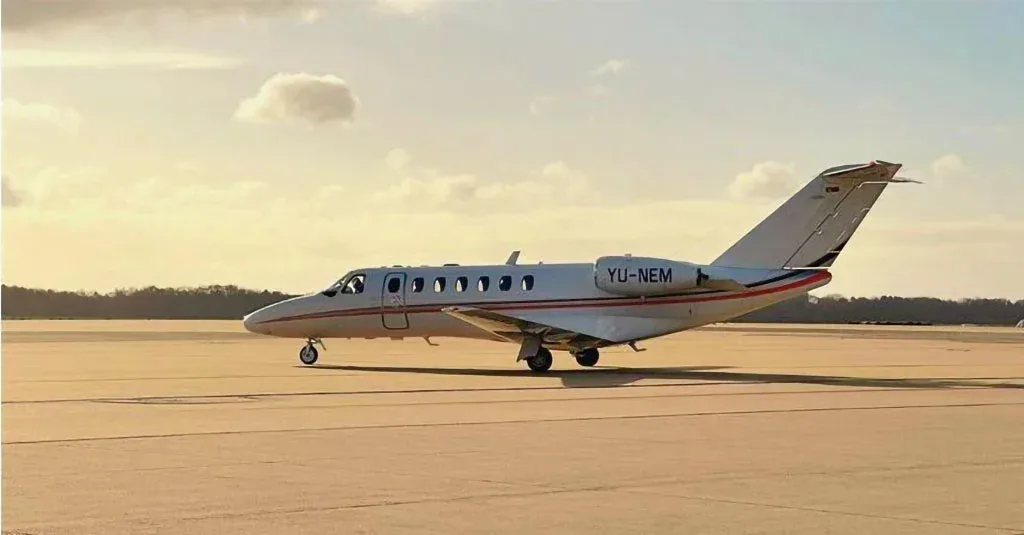 A white Cessna CitationJet with red stripes and registration YU-NEM on an airfield at sunset.