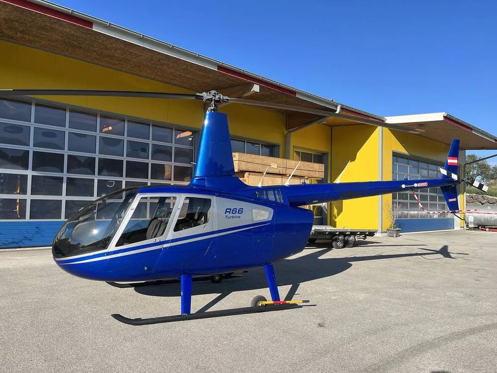 A blue Robinson R66 Turbine helicopter with silver accents and an Austrian flag on the tail, parked on asphalt in front of a yellow hangar.