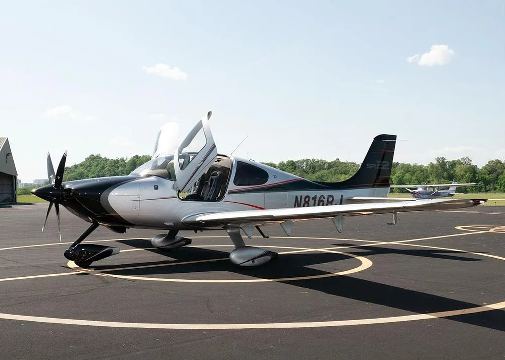 A silver and black Cirrus SR22 aircraft with registration N816RJ is parked on an airfield with its gull-wing doors open, another aircraft visible in the distance.