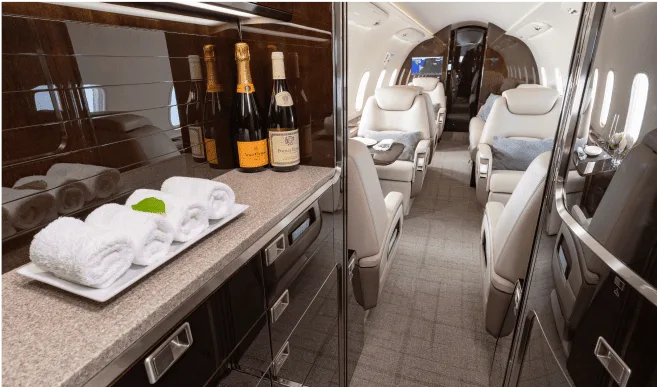 The galley area of a private jet features a counter with champagne bottles and rolled towels, leading into the passenger cabin.