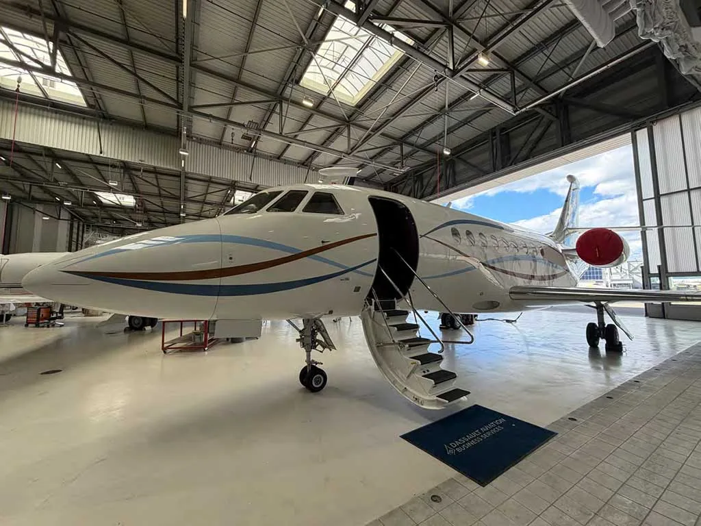 A white business jet with its main door open and stairs extended, parked in a hangar with a "Dassault Aviation" mat on the floor.
