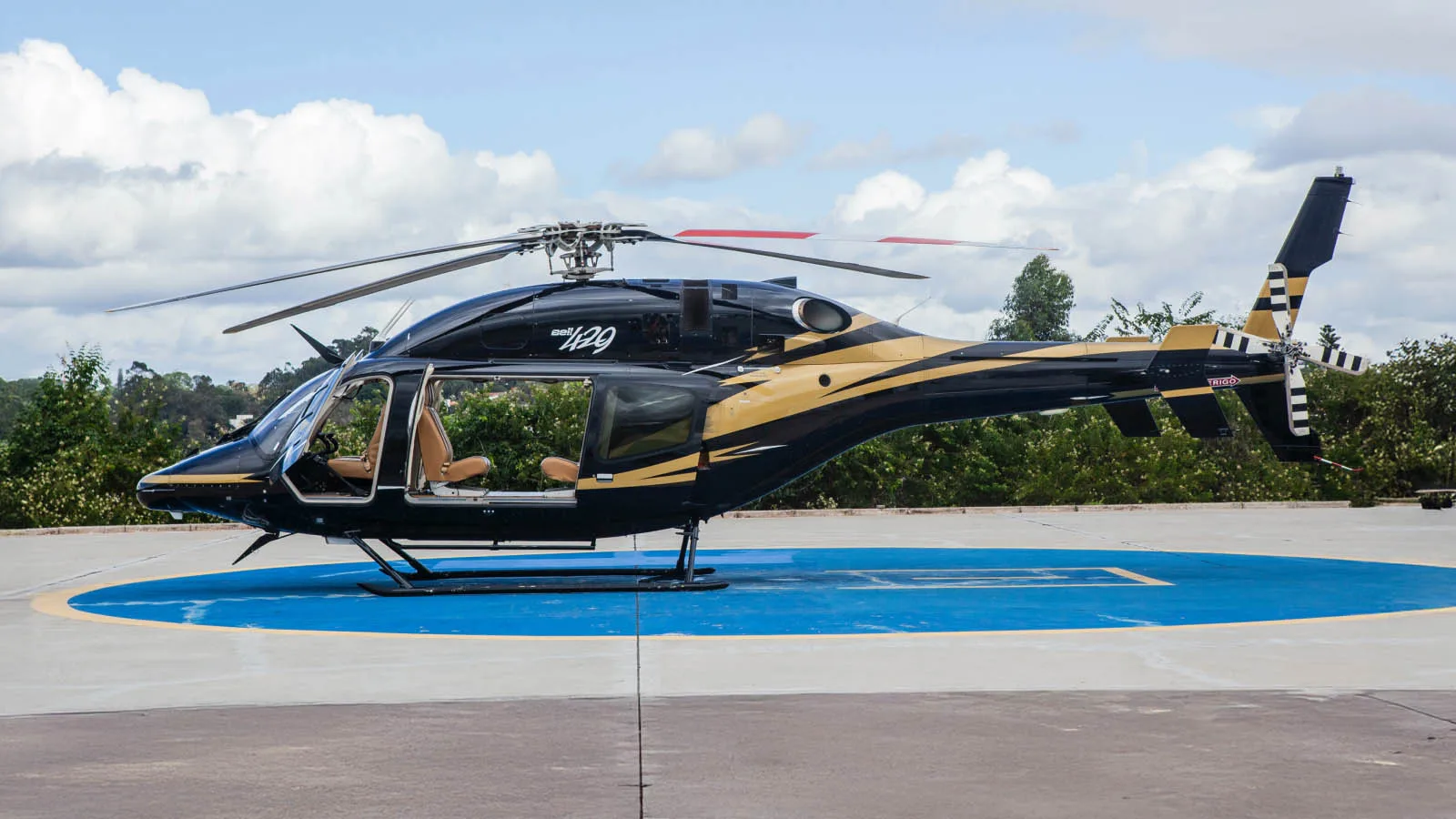 A black and gold Bell 429 helicopter with an open door sits on a blue helipad under a cloudy sky.
