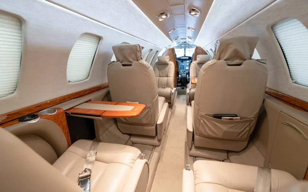 A view down the aisle of a private jet cabin with beige leather seats and wood trim, looking towards the cockpit.