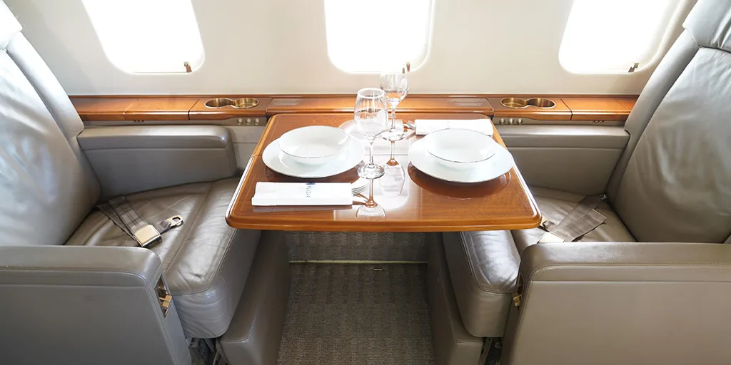 A private jet cabin interior showing two light grey leather seats facing each other with a polished wooden table set with plates and glasses between them.