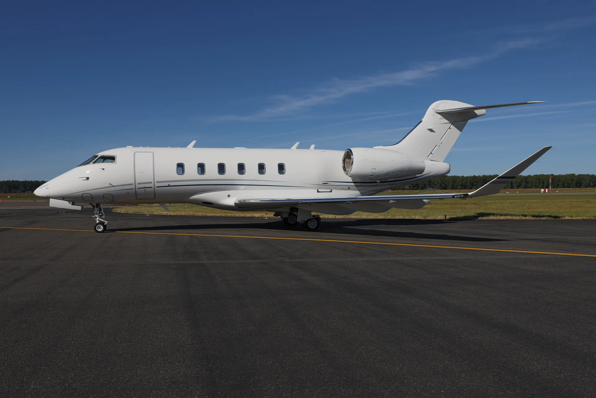 A white business jet with blue and grey accent stripes is parked on an asphalt runway under a clear blue sky.
