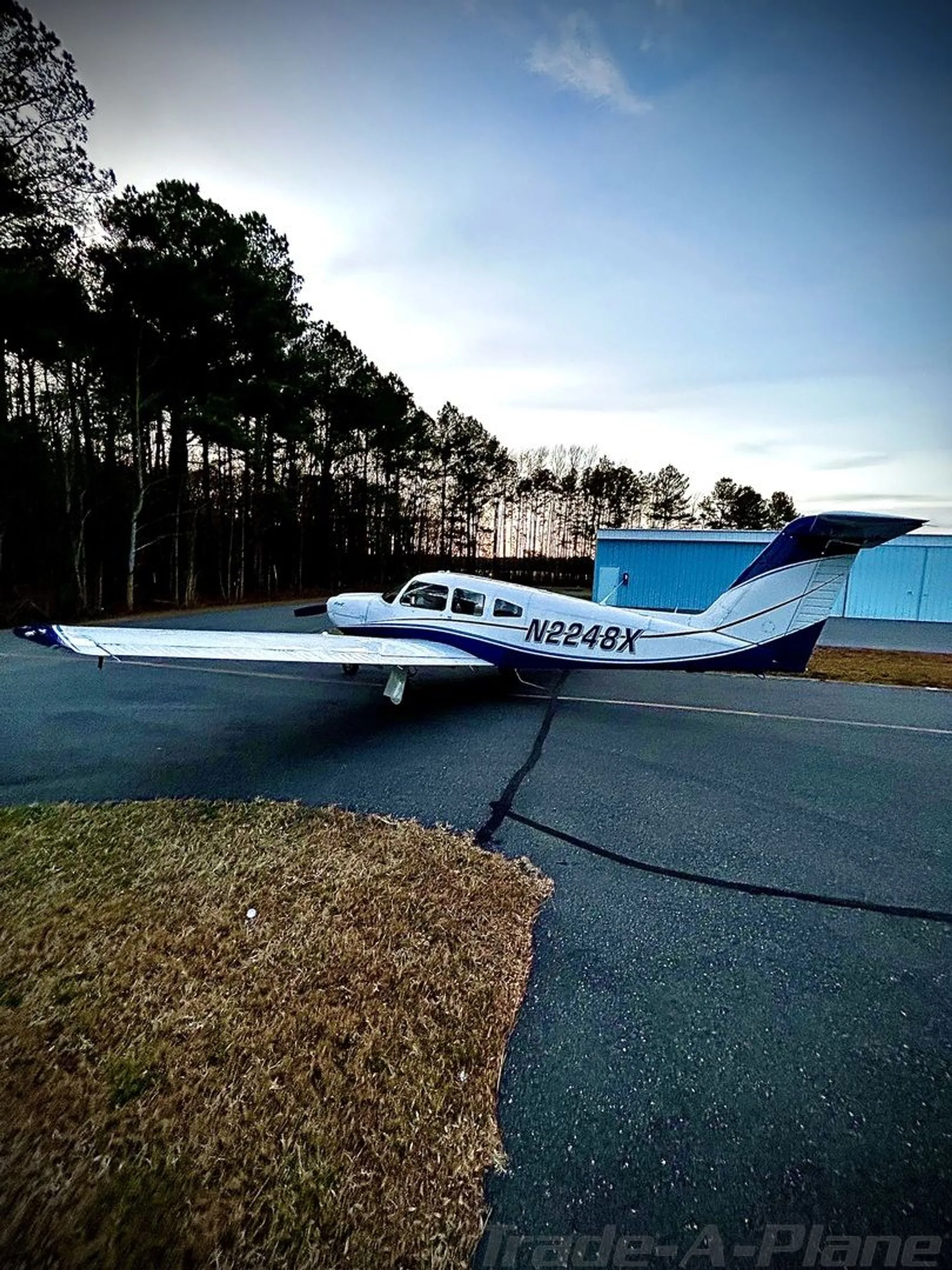 A white and blue Piper Archer II (N2248X) parked on an asphalt ramp near trees at dusk.