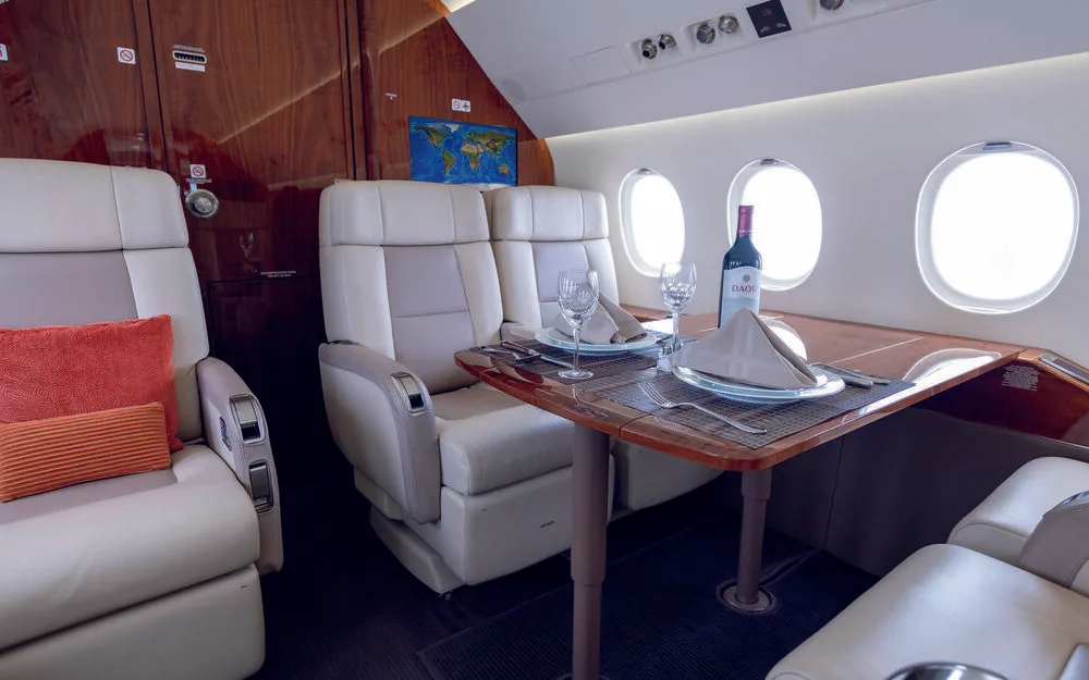 A close-up view of the business jet's cabin dining area, featuring beige seats, a wooden table set with wine, glasses, and place settings, next to large windows.
