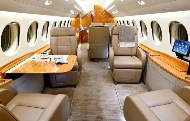 A private jet cabin interior with beige leather seats, a wooden table set with a coffee cup, and a digital display.