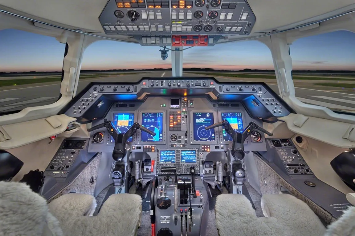 The modern cockpit of a private jet with multiple digital displays and controls, overlooking a runway at dusk.