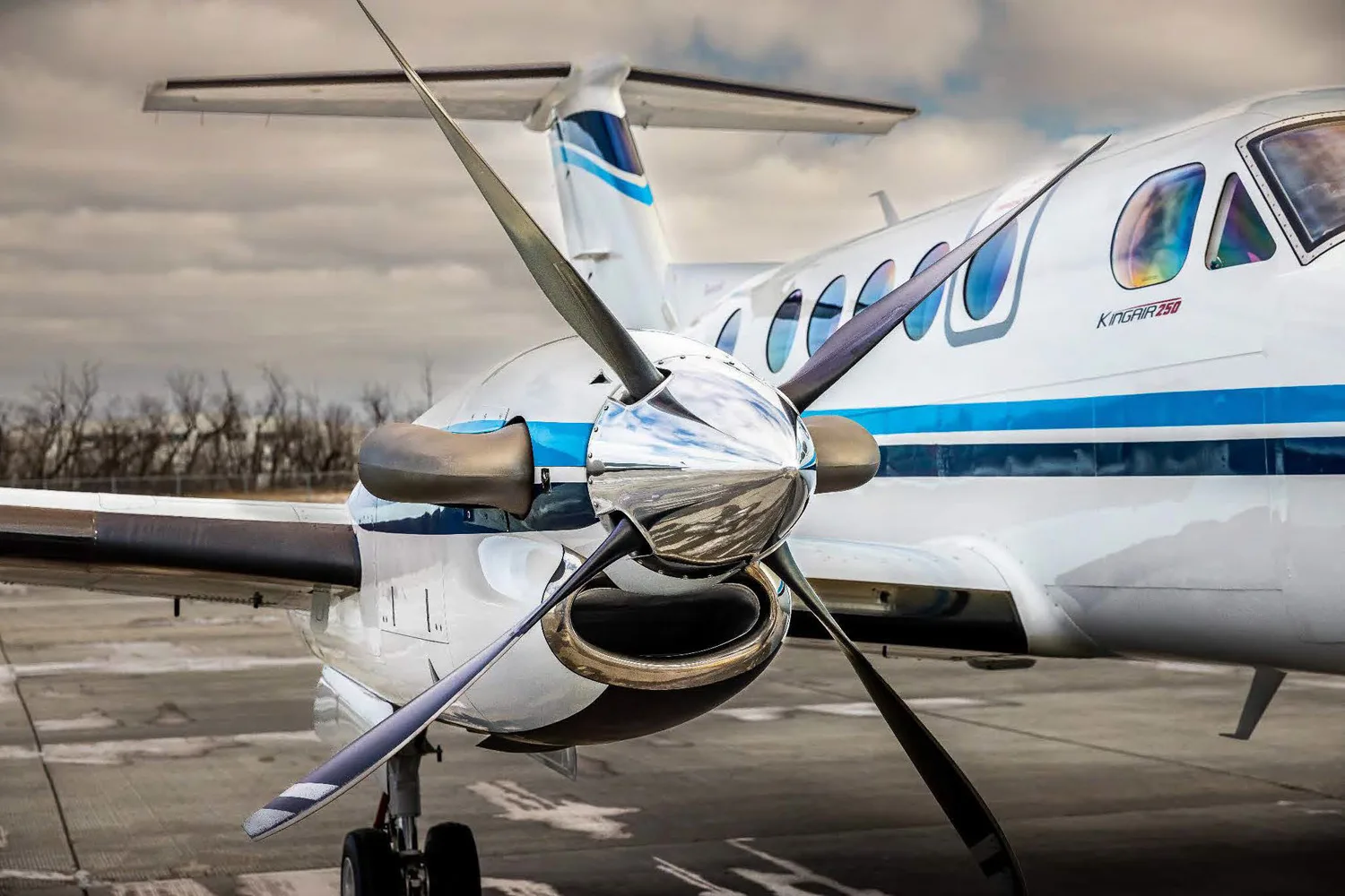 A close-up view of the propeller and front fuselage of a white King Air 250 turboprop aircraft with blue stripes on an airport tarmac.