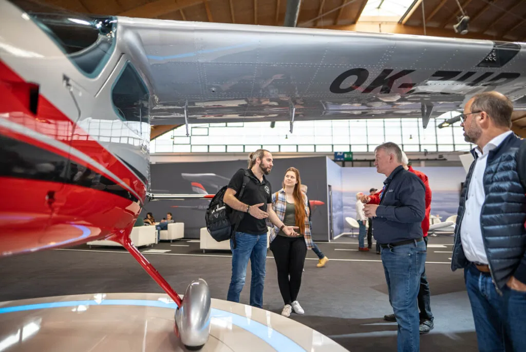 A red and silver light aircraft with registration OK-ZUB on display at an indoor exhibition, with people standing nearby.