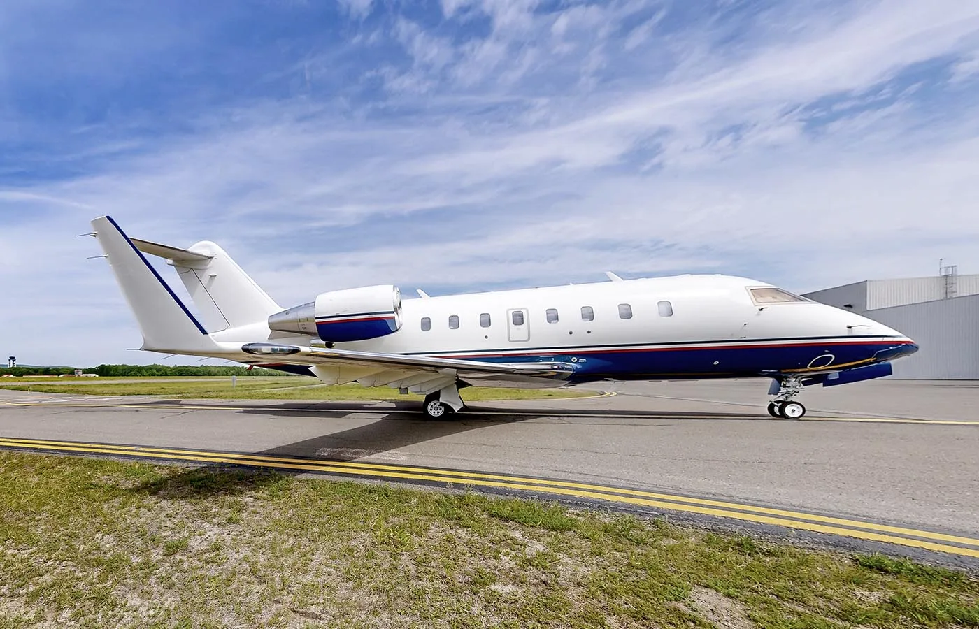 A white and blue Bombardier Challenger business jet is parked on a tarmac under a partly cloudy sky.