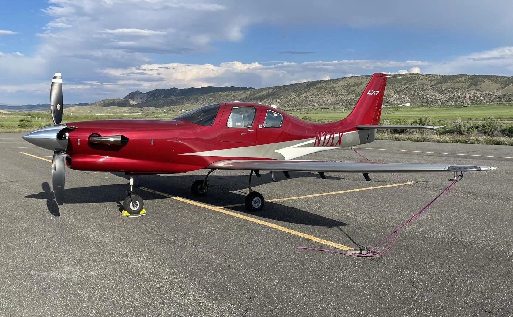 A red and silver Lancair LX7 single-engine propeller aircraft (N17LX) is parked on an asphalt runway with mountains in the background under a partly cloudy sky.