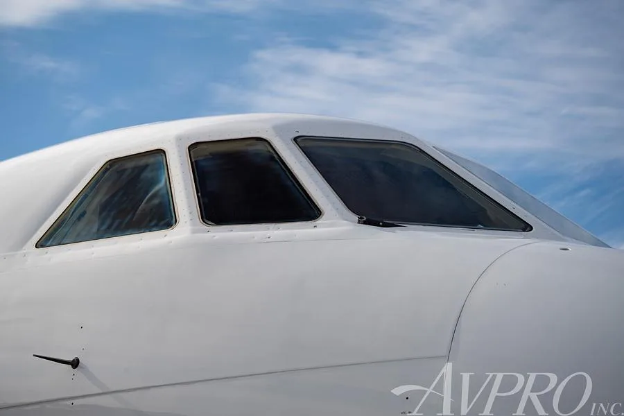 A close-up view of the cockpit windows of a white Dassault Falcon business jet under a blue sky.