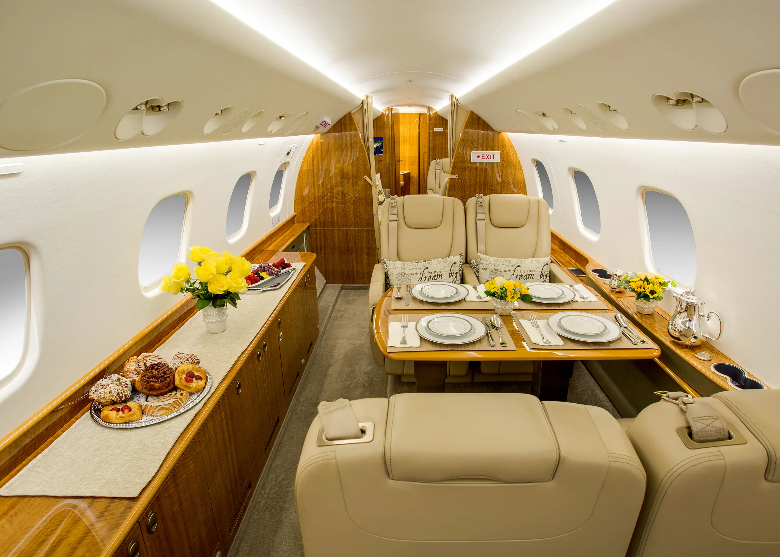 A wide view of a private jet's elegant cabin interior, featuring beige seating, a dining table, and a buffet counter with food.