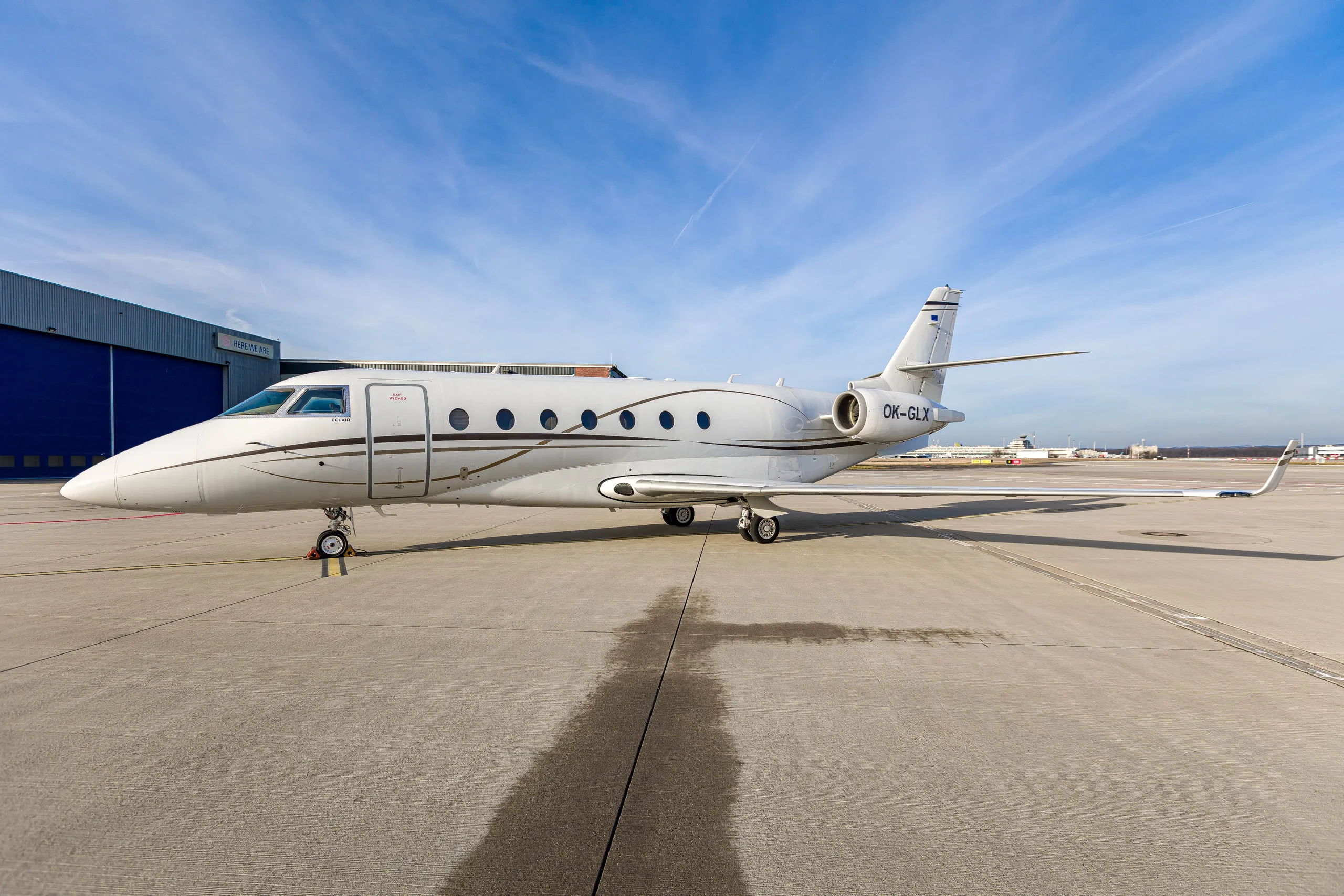 A white Gulfstream G200 business jet with registration OK-GLX parked on an airport tarmac under a blue sky.