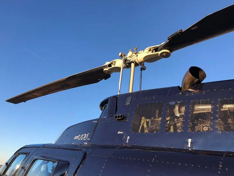 A close-up view of the main rotor head and exhaust of a dark blue Bell 206 helicopter against a clear blue sky.