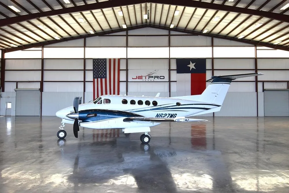 A white and blue Beechcraft King Air twin-engine turboprop aircraft, N627MG, is parked in a hangar with US and Texas flags.