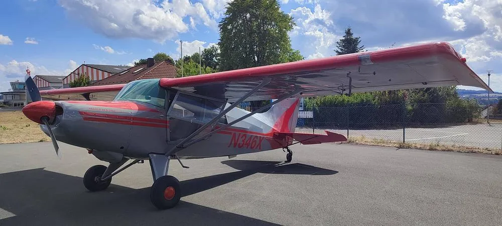 A gray and red single-engine aircraft, N346X, parked on an asphalt apron under a partly cloudy sky.
